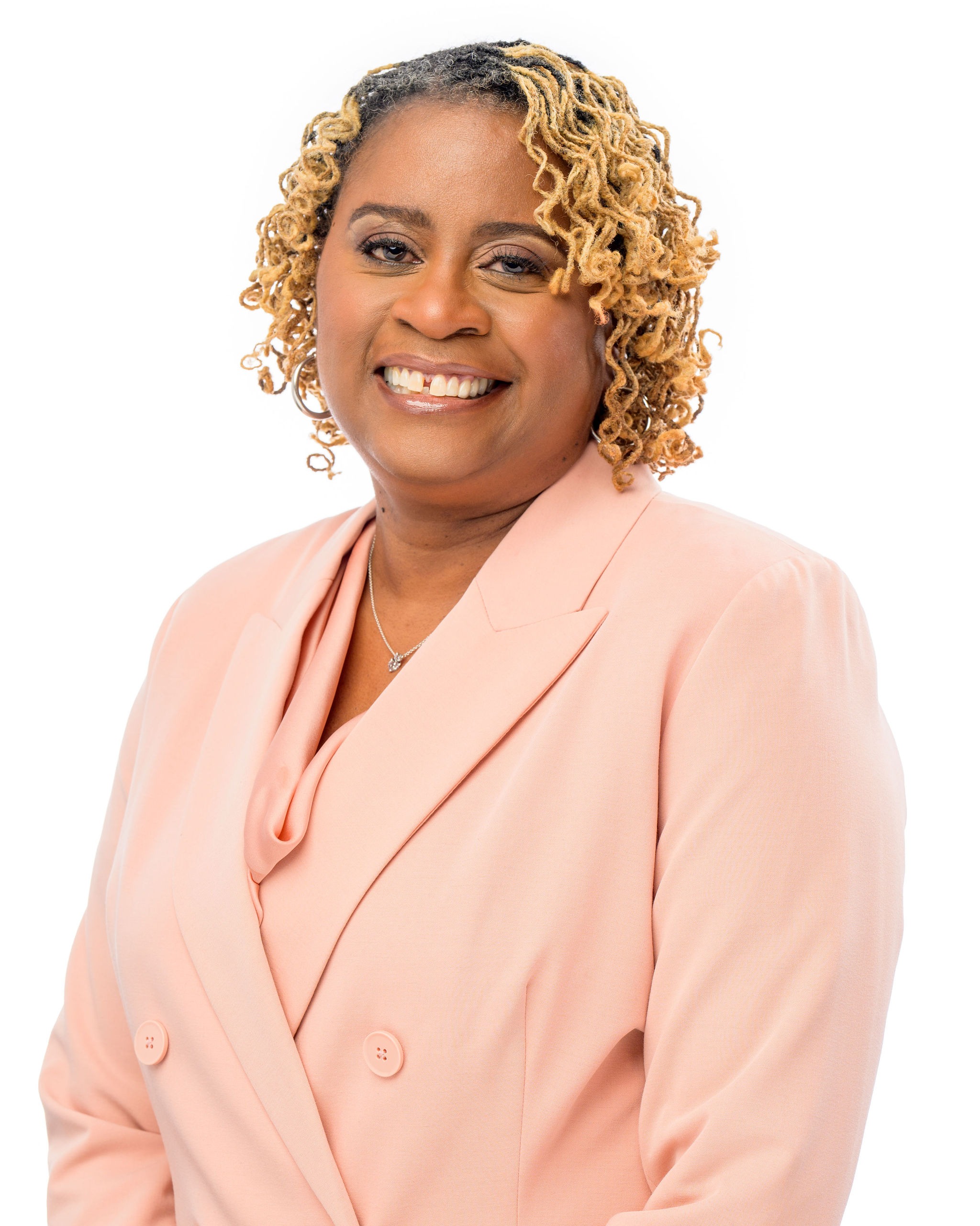 A woman with short, curly blonde hair smiles while wearing a light pink blazer over a matching top, standing against a plain white background.