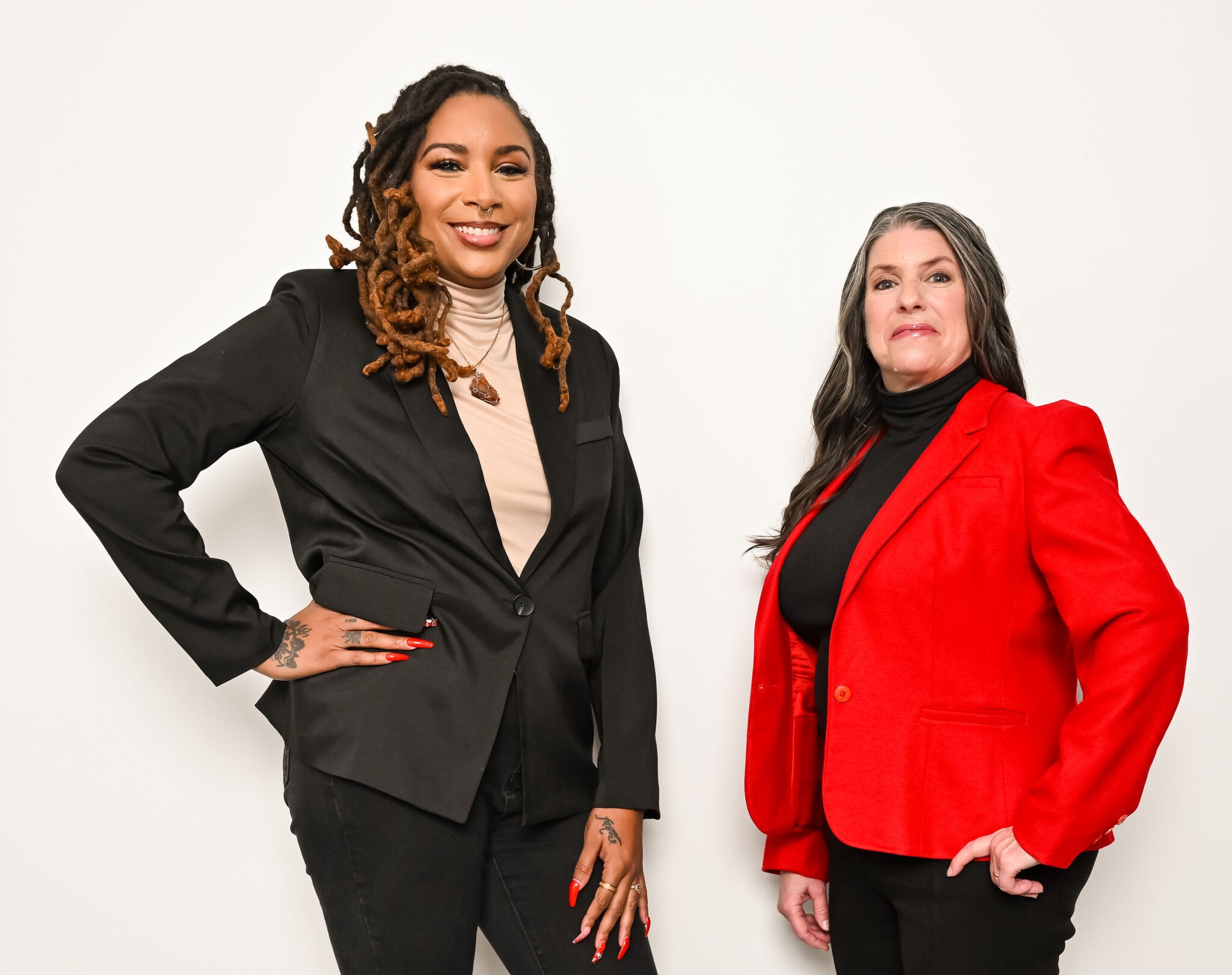 Two women stand side by side against a white background. One wears a black blazer and beige top with long, braided hair. The other wears a red blazer and black top with long, straight hair. Both are smiling confidently.