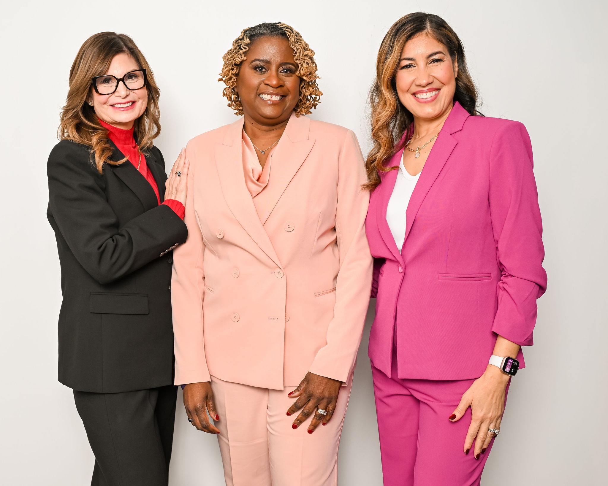Three women in business suits stand smiling against a plain white background. The woman on the left wears a black suit, the middle wears a light pink suit, and the right wears a bright pink suit.