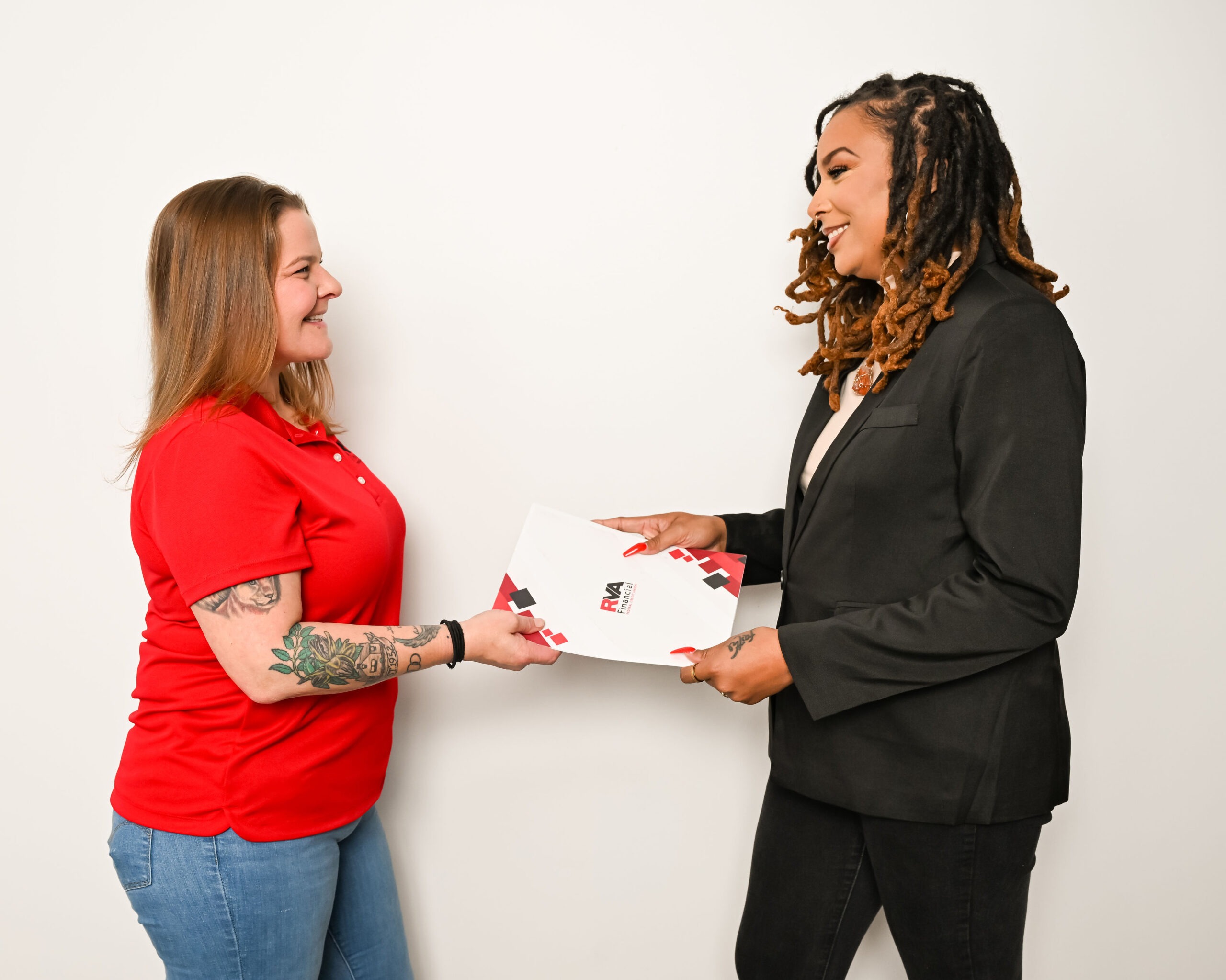Two women standing and smiling at each other; one in a red shirt and jeans hands a folder to the other, who is wearing a black blazer. The folder has a red and black design.
