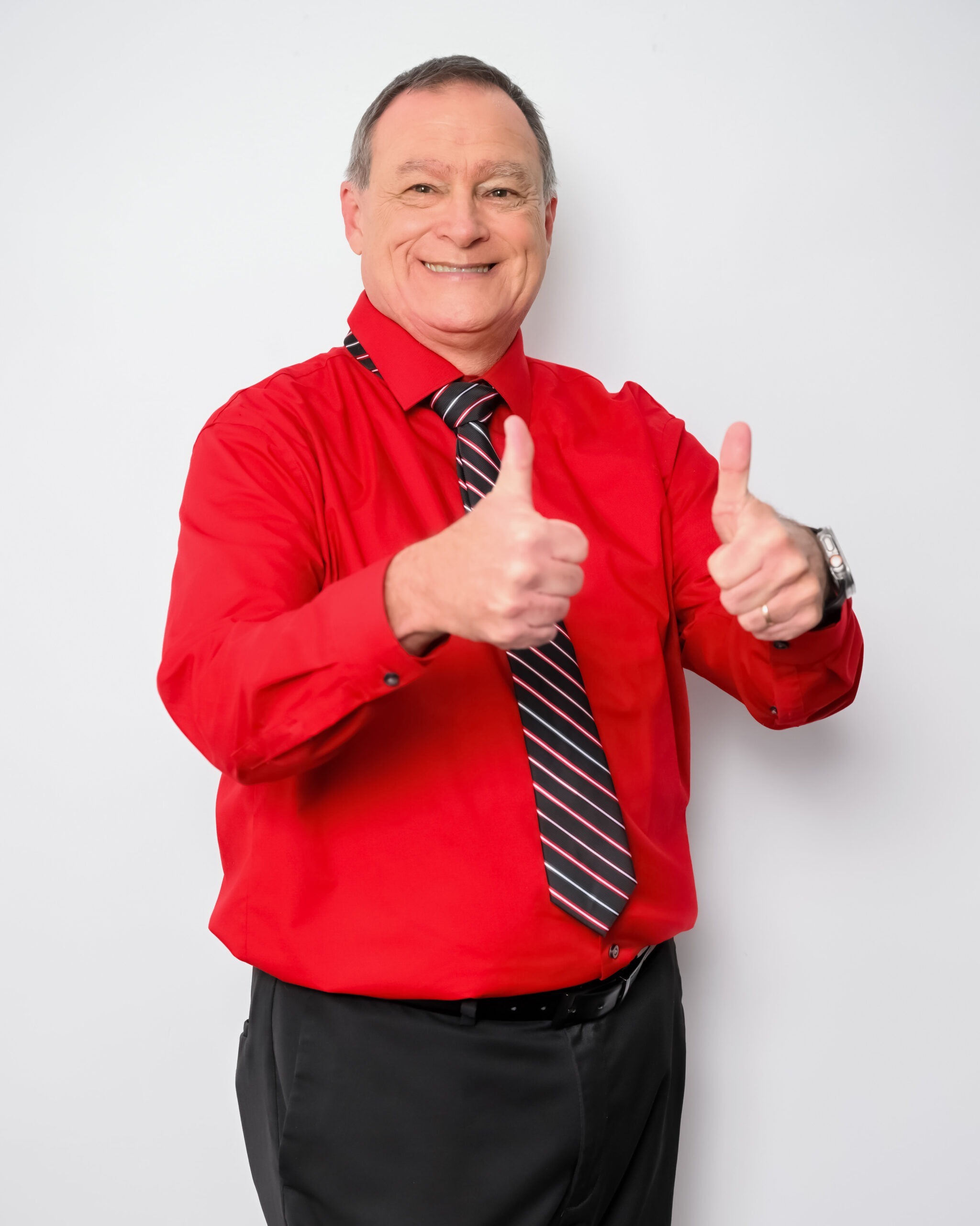 Smiling man wearing a bright red shirt and striped tie gives two thumbs up while standing against a plain white background.