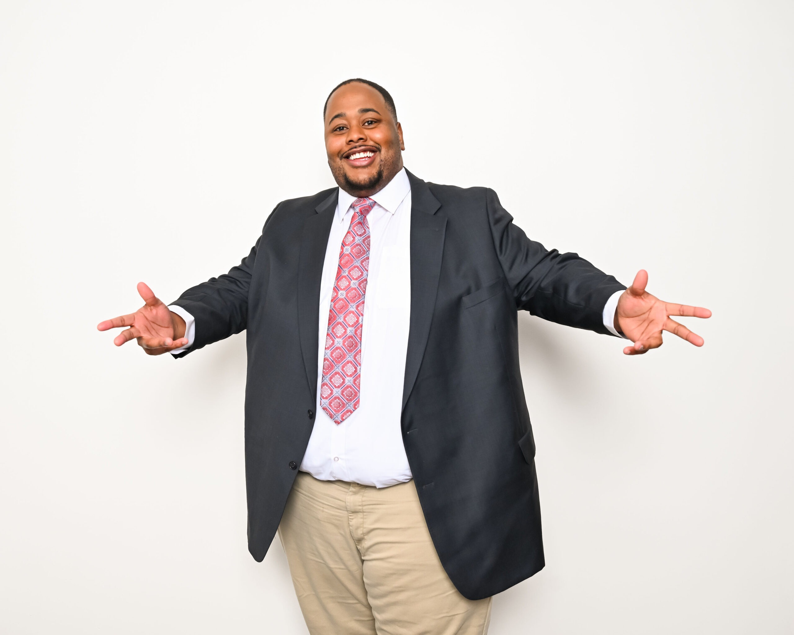 Smiling Black man wearing a black suit, white shirt, and patterned red tie, stands with arms outstretched in a welcoming gesture against a white backdrop.