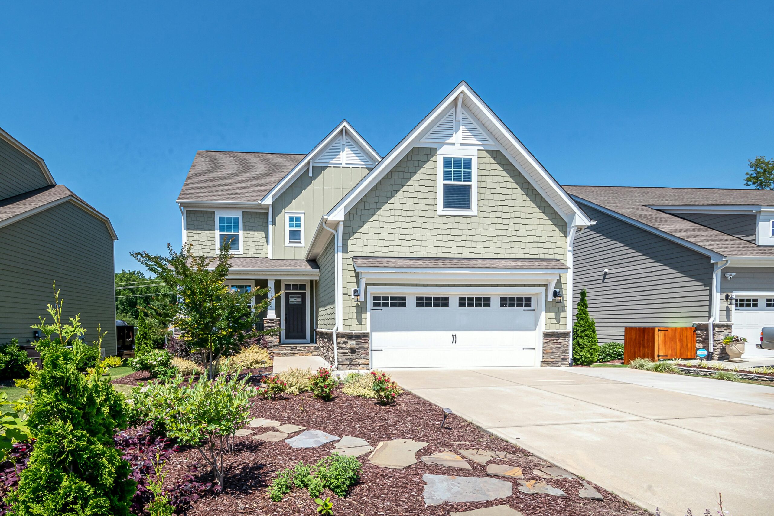 A modern two-story suburban house with light green siding, white trim, a double garage, and a neatly landscaped front yard under a clear blue sky.