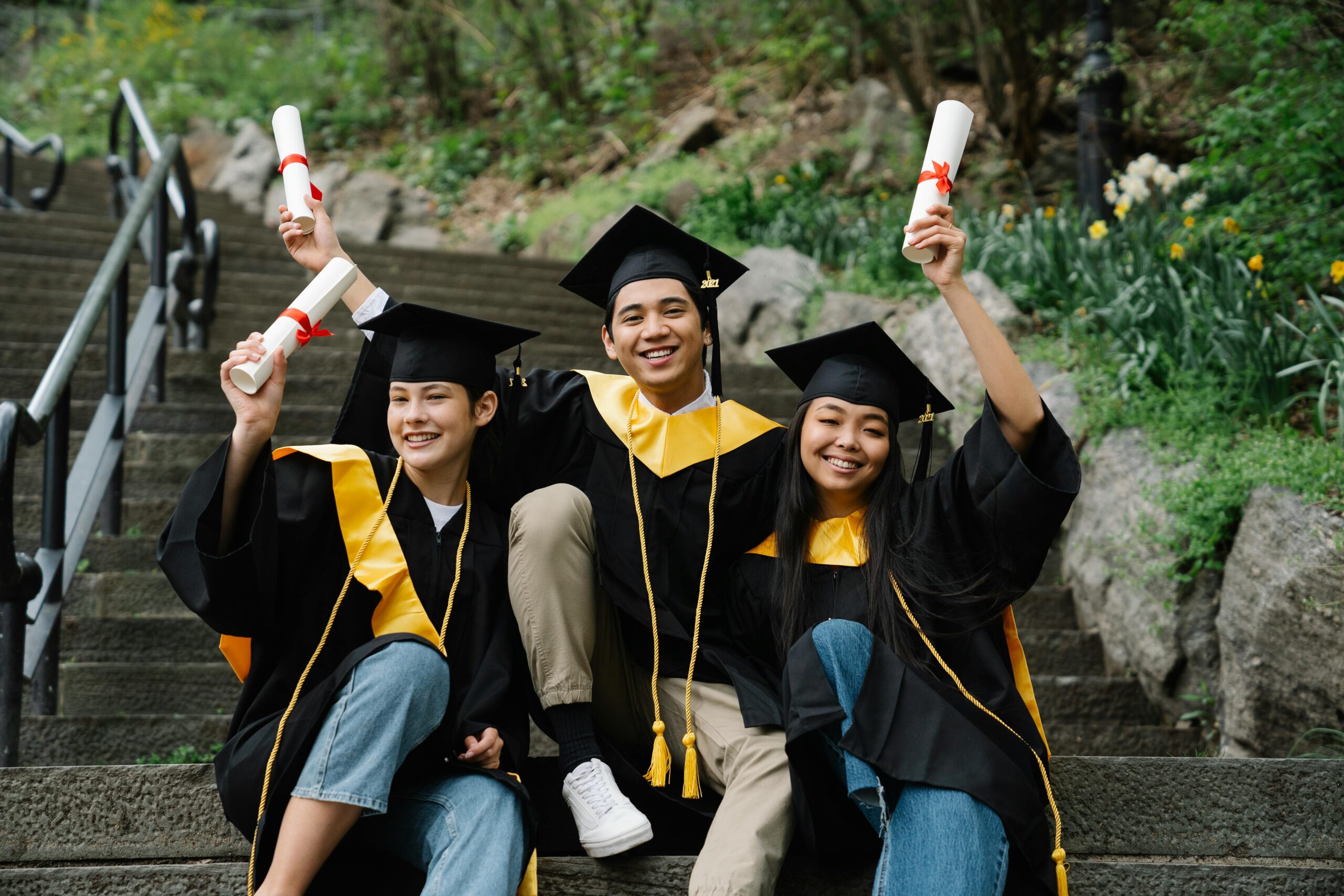 Three graduates in caps and gowns sit on outdoor steps, smiling and holding up their diplomas. Trees and greenery are in the background, creating a celebratory and joyful atmosphere.
