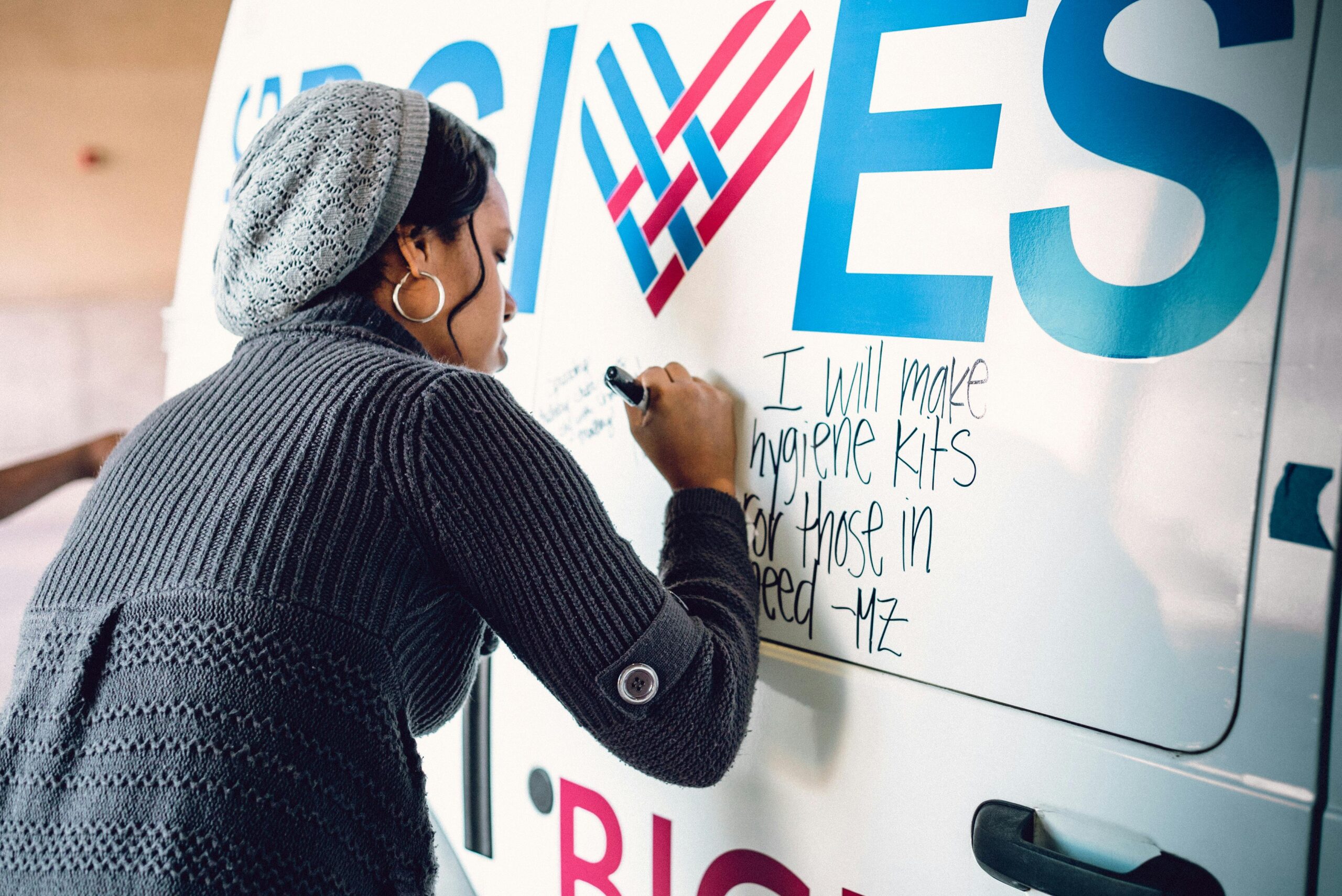 A woman in a gray knit hat and sweater writes a pledge on a van, stating, I will make hygiene kits for those in need. The van features a large heart in its design.