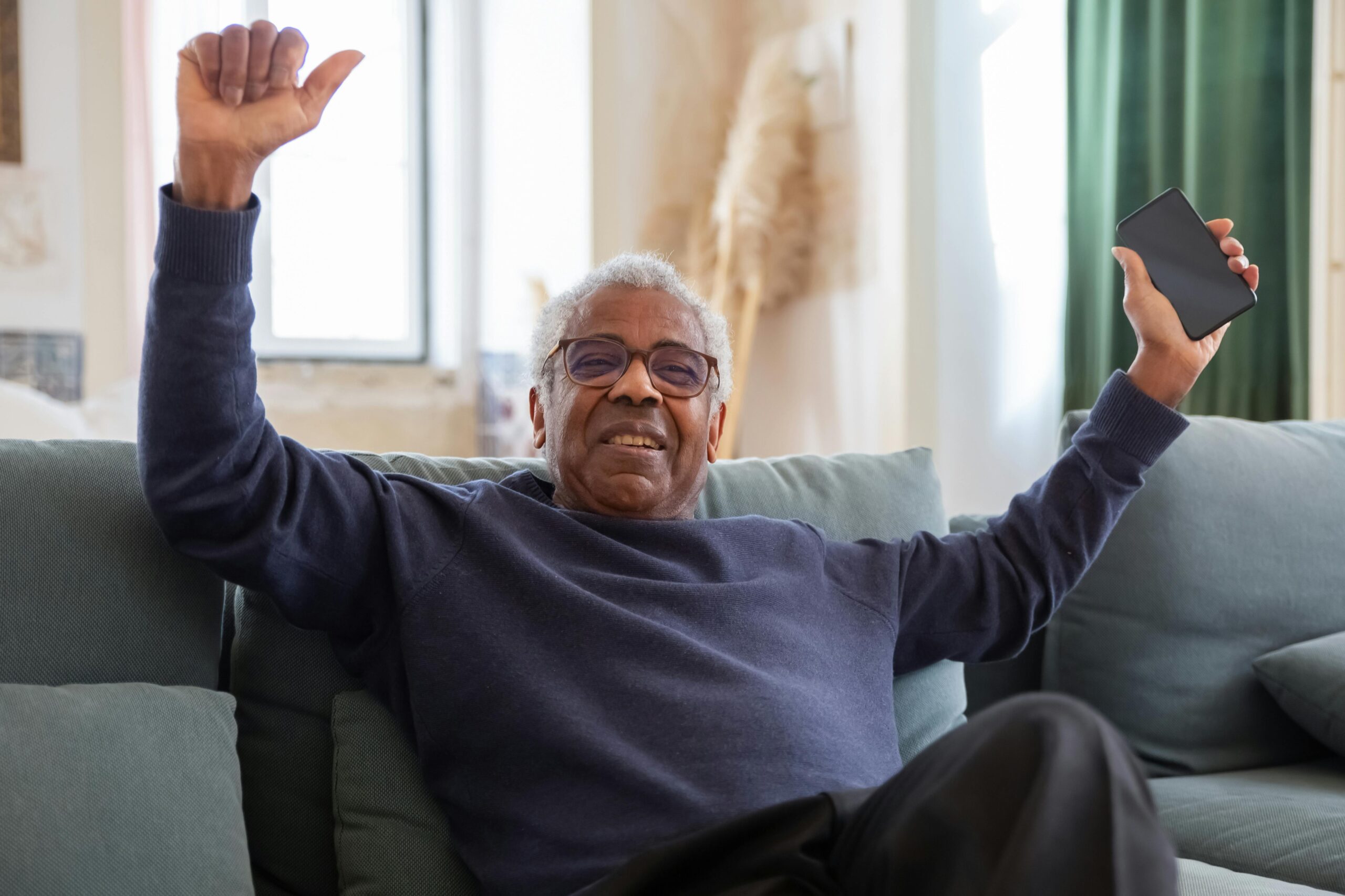An older man with gray hair wearing glasses and a navy sweater sits on a couch, smiling and raising his arms in celebration while holding a smartphone in one hand.