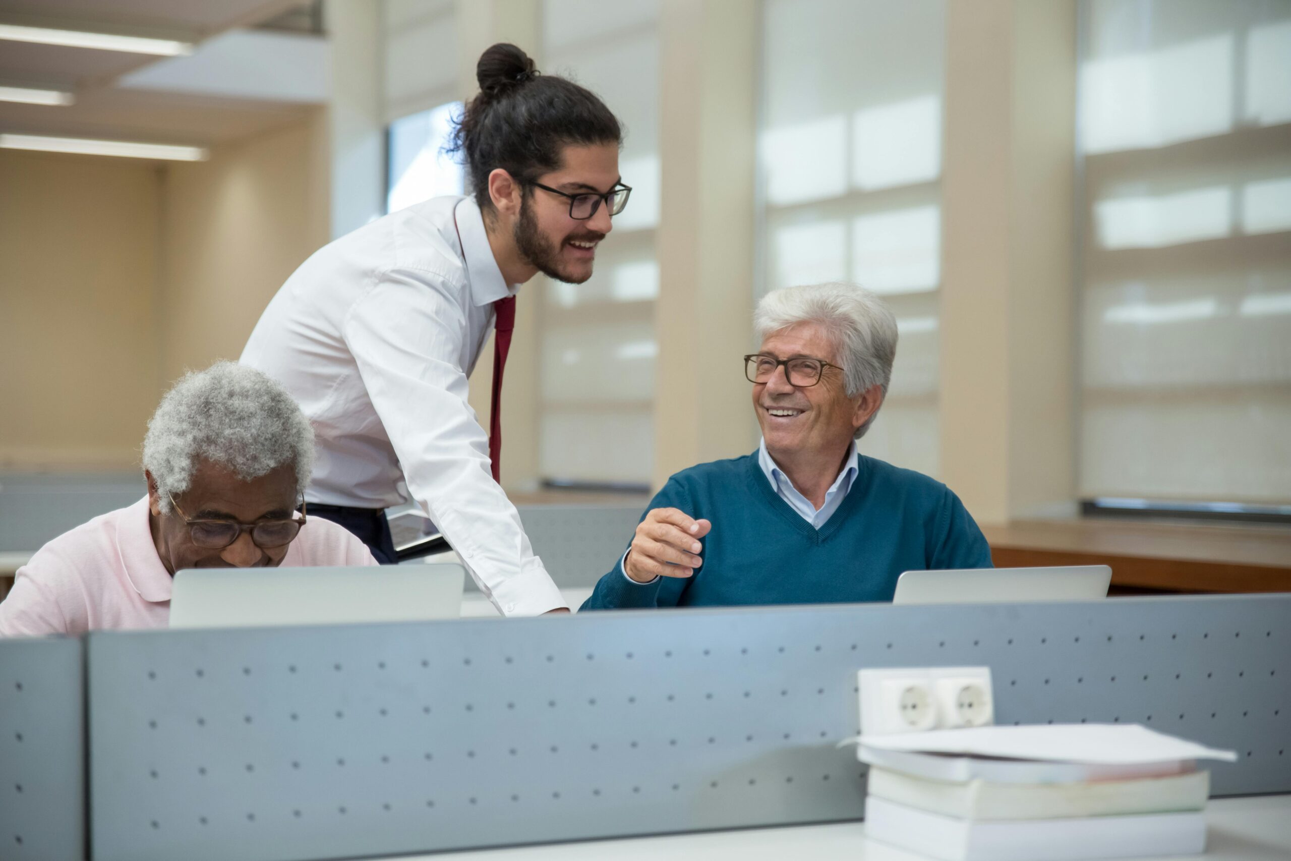 A young man with a bun, in a white shirt and tie, assisting an elderly man with gray hair, wearing glasses and a blue sweater, in a computer lab setting. An older black man sits at a computer at the left.