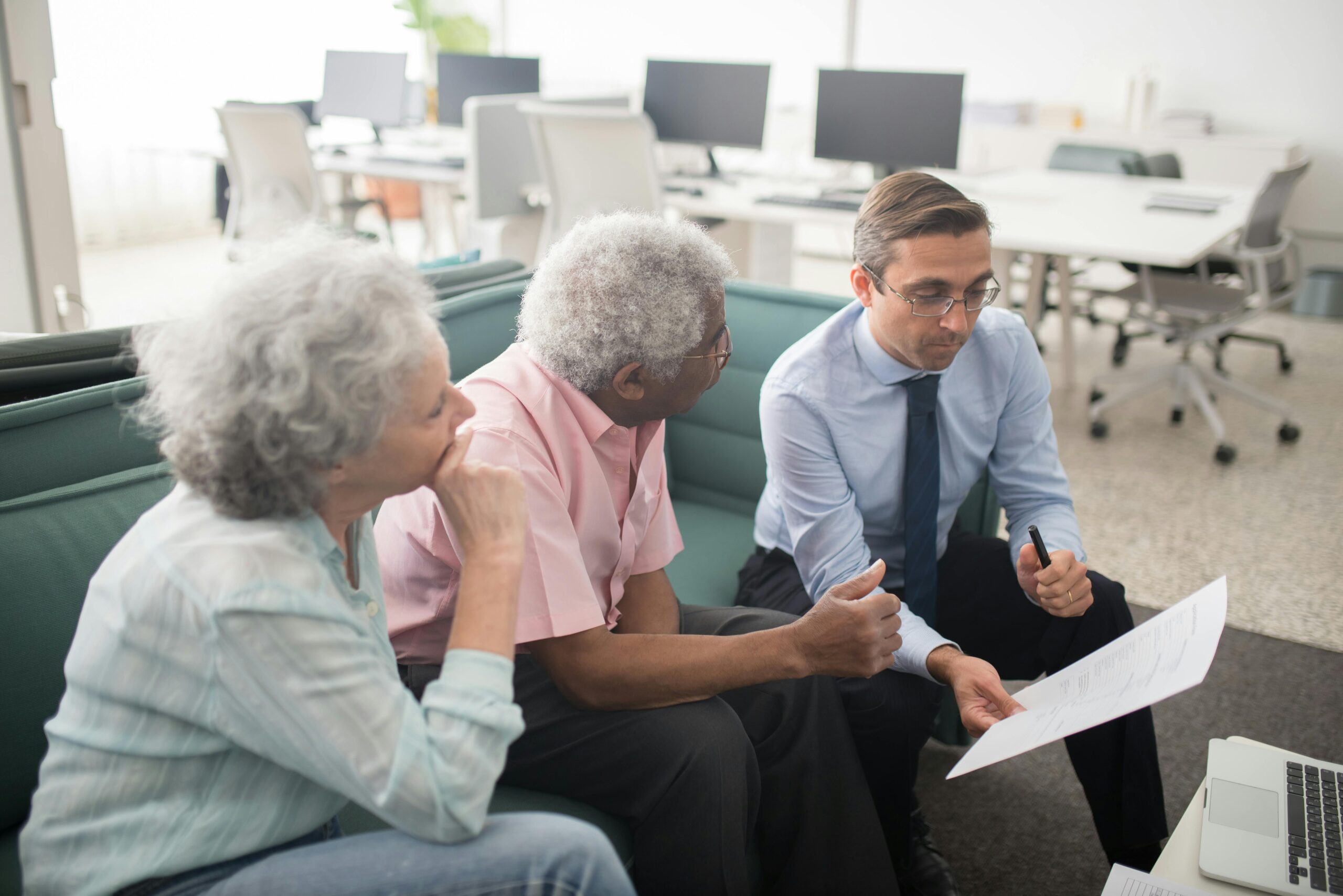 A man in business attire shows documents to two older adults sitting on a couch in a modern office, discussing paperwork together.