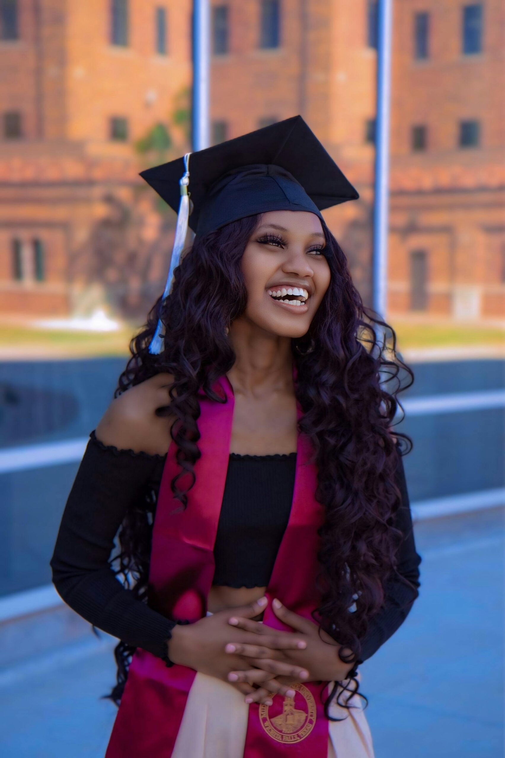 A young woman wearing a graduation cap, burgundy stole, and black off-shoulder top smiles joyfully while standing outdoors in front of a brick building.