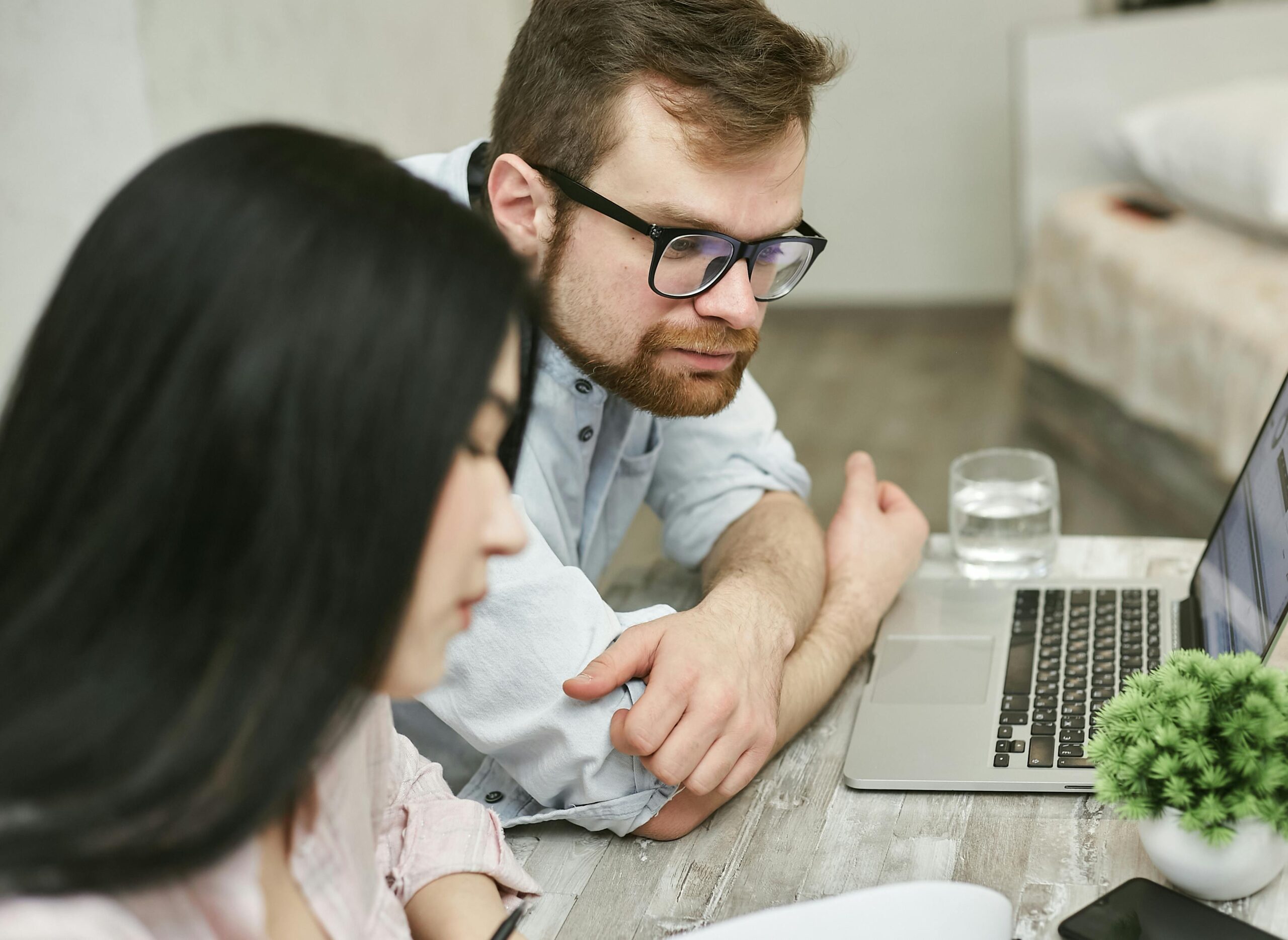 A man with glasses and a beard looks at a laptop screen while sitting at a wooden table beside a woman with long dark hair. A glass of water and a small potted plant are also on the table.