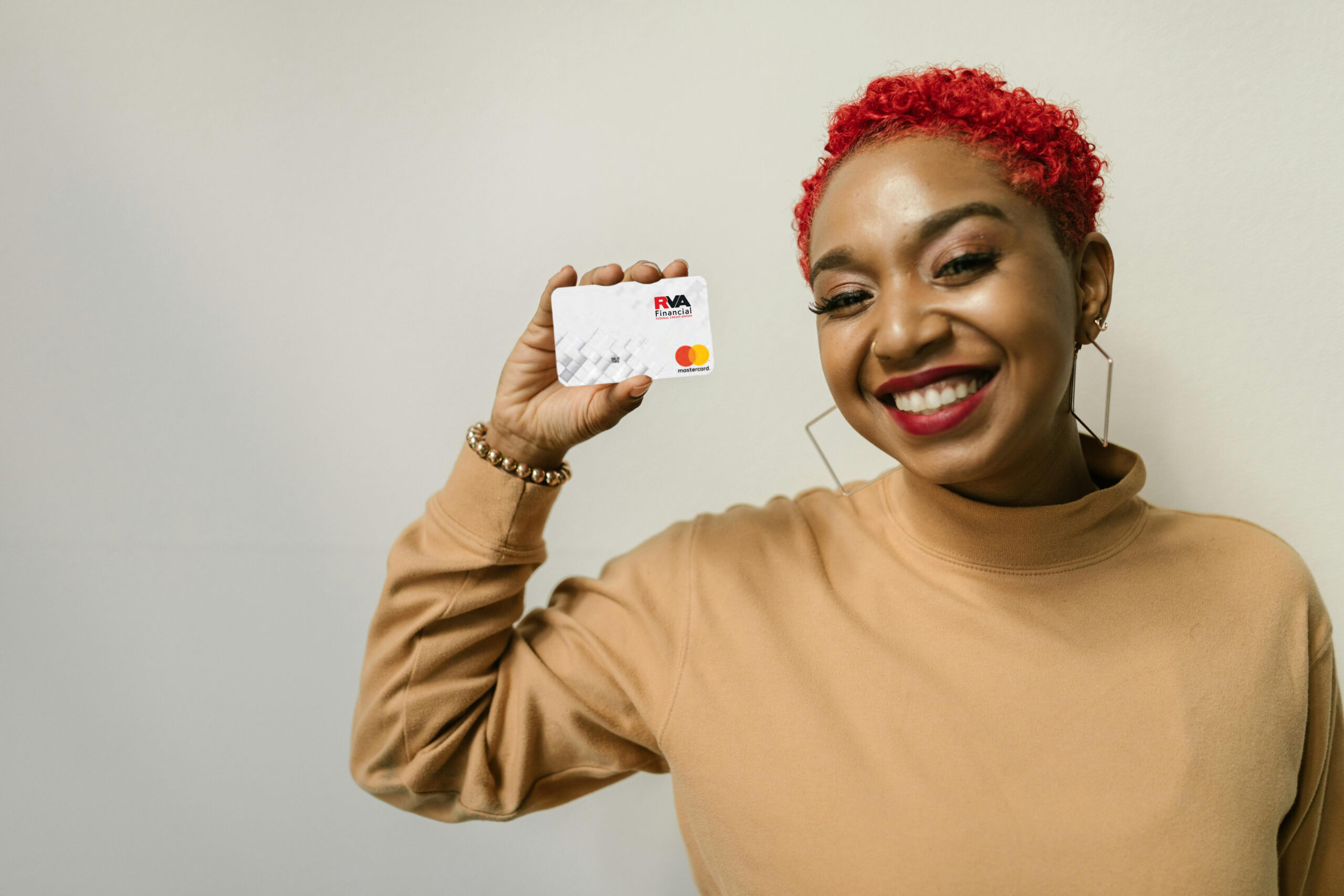 A smiling woman with short curly red hair holds up a white credit card with a Mastercard logo. She wears a tan turtleneck and hoop earrings, standing against a plain light background.