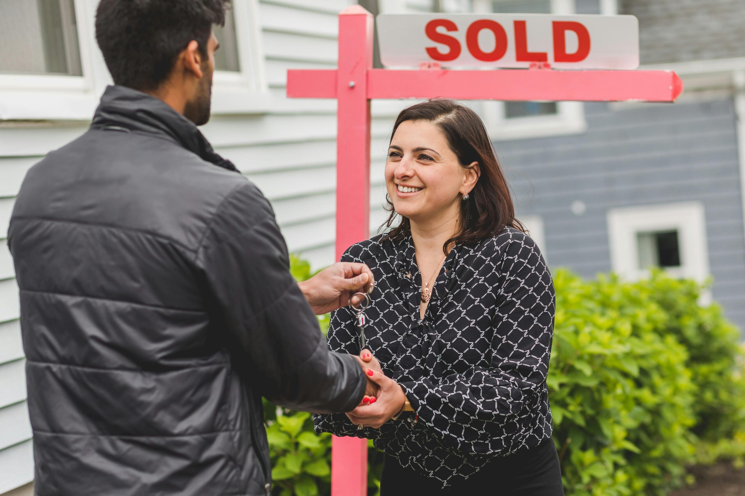 A smiling woman, likely a realtor, receives keys from a man as a "SOLD" sign sits nearby in front of a house.