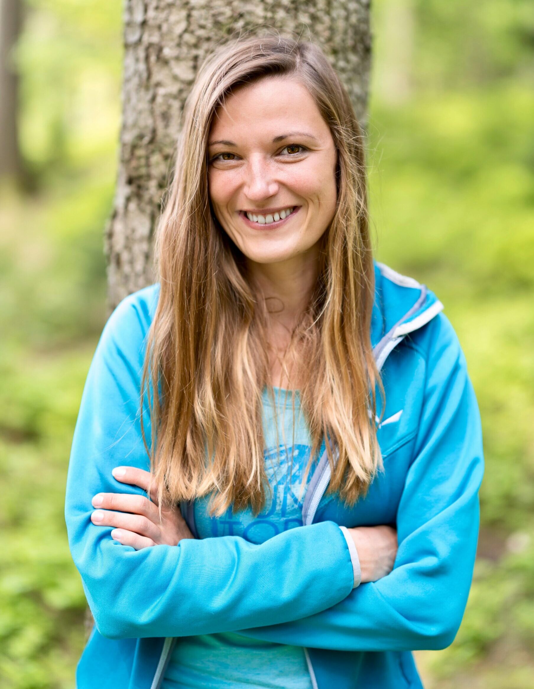 A woman with long blonde hair and a bright smile, wearing a blue zip-up jacket, stands in a forest with her arms crossed.