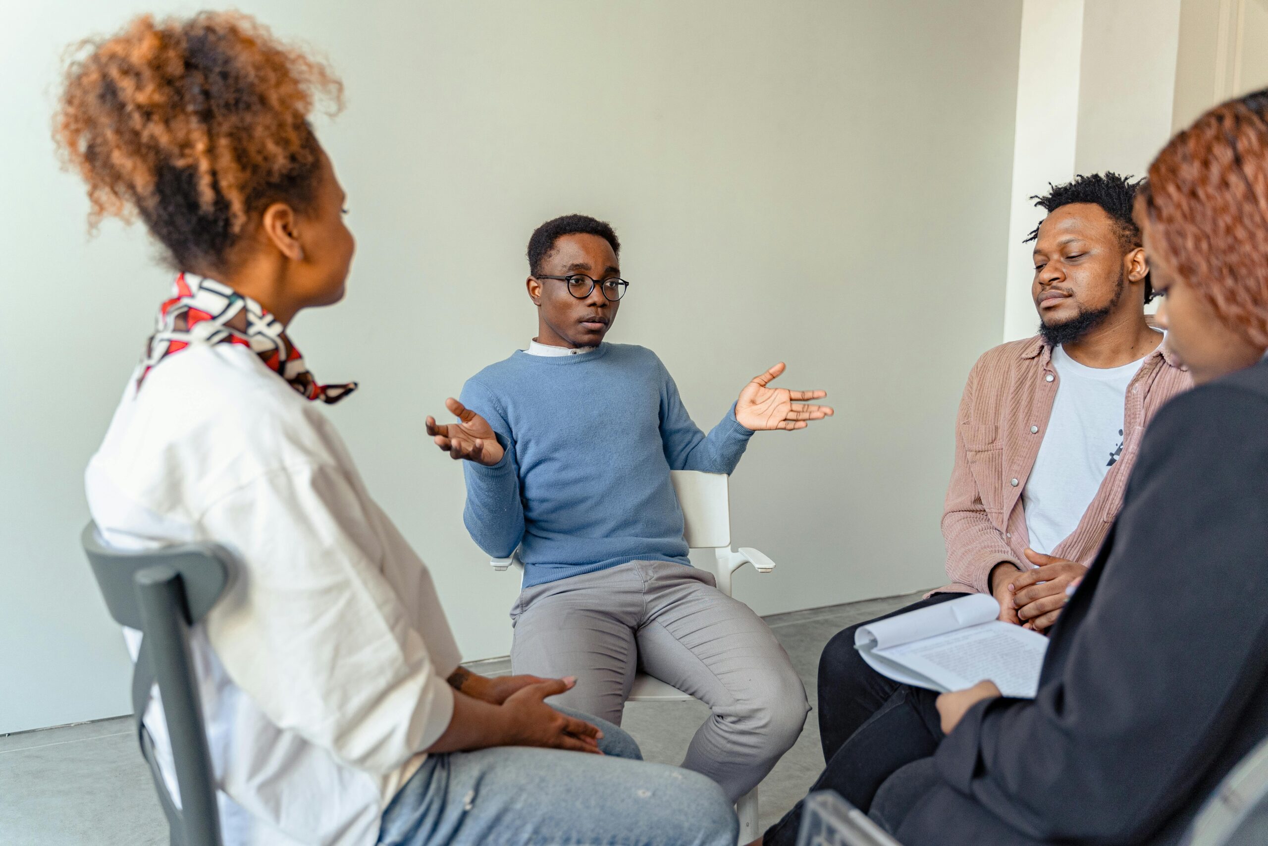 Four people sit in a circle having a discussion. One man in a blue sweater gestures with his hands while the others listen attentively, creating an atmosphere of group conversation or counseling.