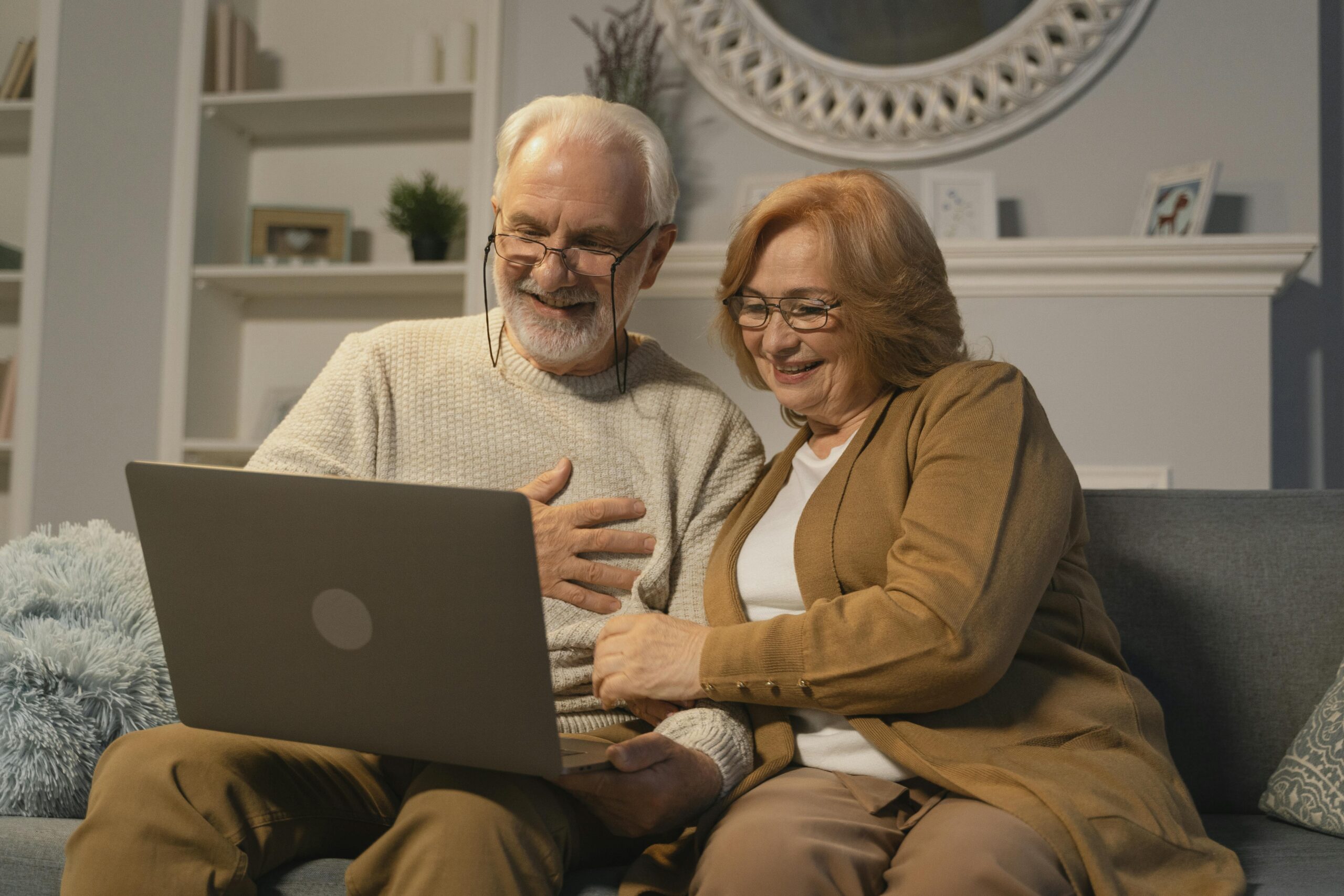An elderly couple sits on a couch, smiling and looking at a laptop together in a cozy living room. They appear happy and engaged, sharing a pleasant moment.