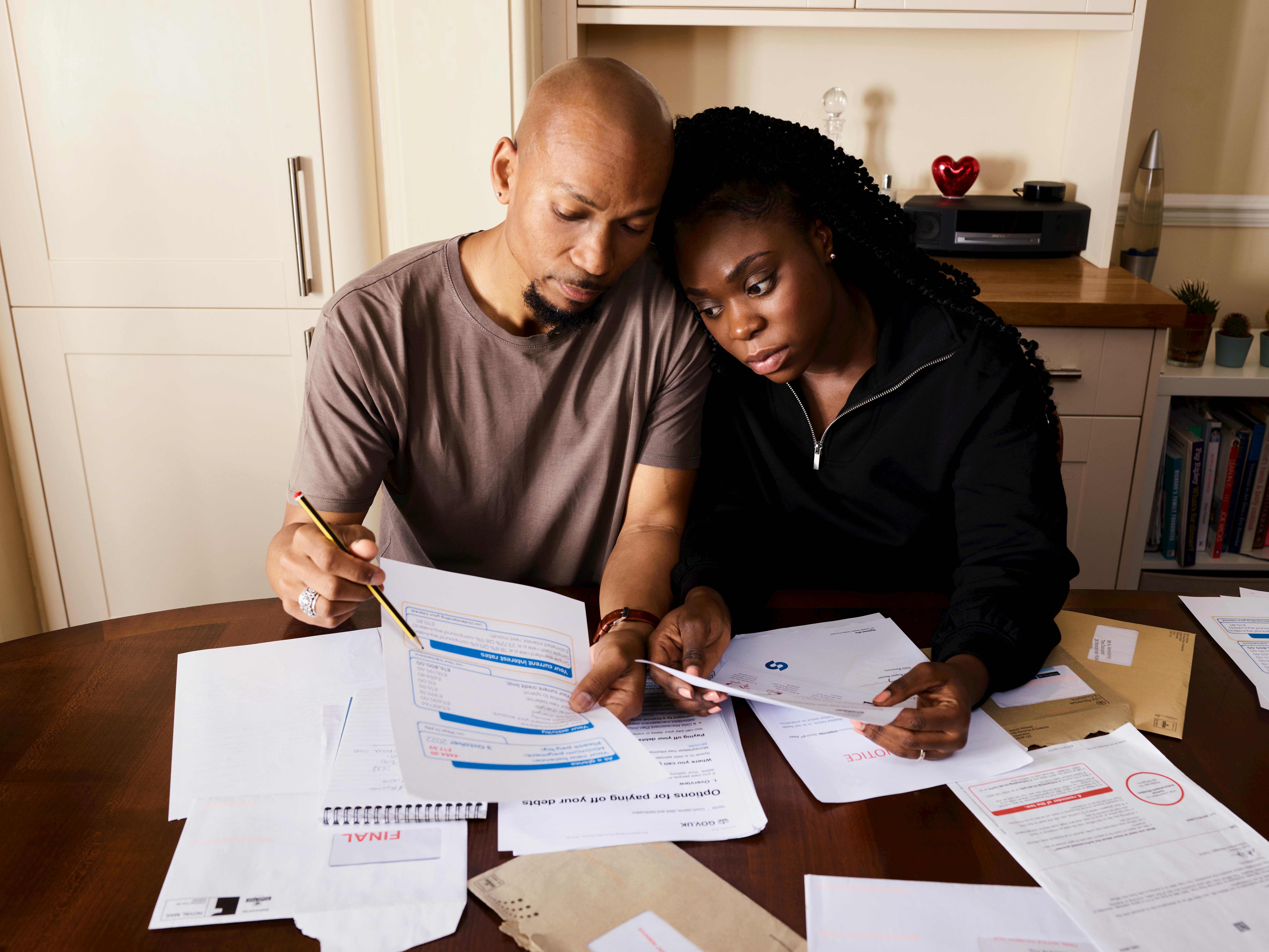 An African-American couple, a man in a brown shirt and a woman in a black jacket, sits at a wooden table reviewing financial paperwork, the man pointing at a document with a pencil, while the woman looks on with a concerned expression.